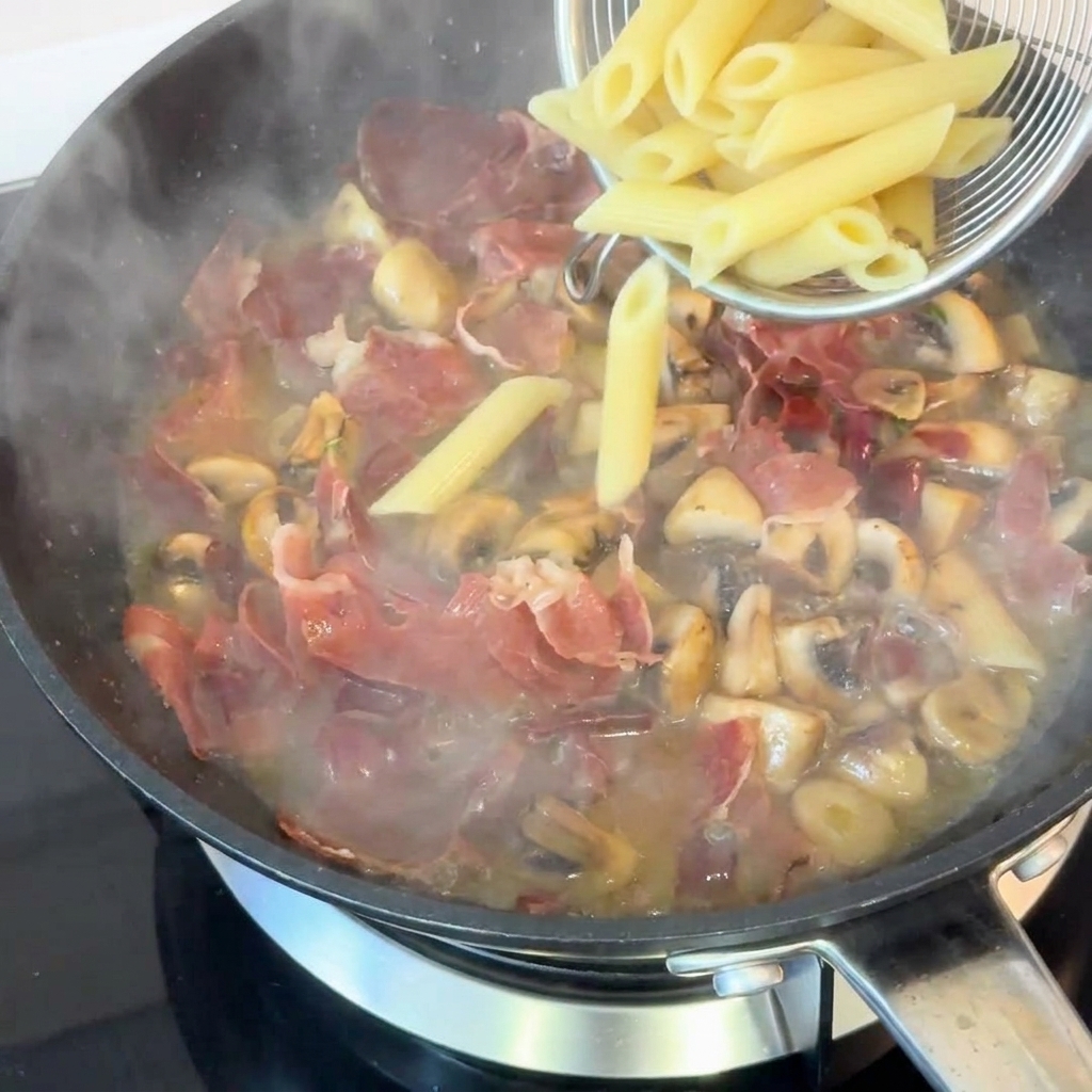 Pennes cuites récupérées avec une écumoire et déposées dans une poêle pleine de jambon savoureux, de champignons et de sauce riche.