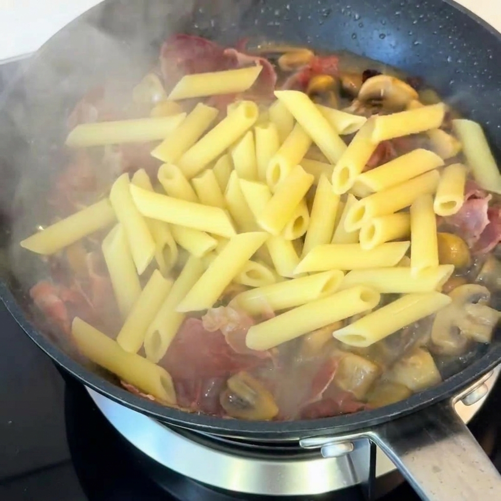 Pennes cuites mélangées dans une poêle avec des champignons sautés et du jambon, assaisonnées de poivre noir.