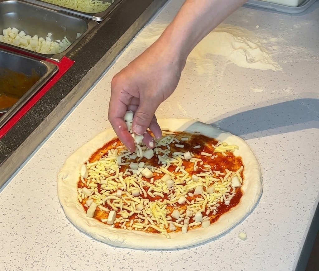 Une main saupoudrant du fromage râpé blanc sur une croûte à pizza garnie de sauce tomate.