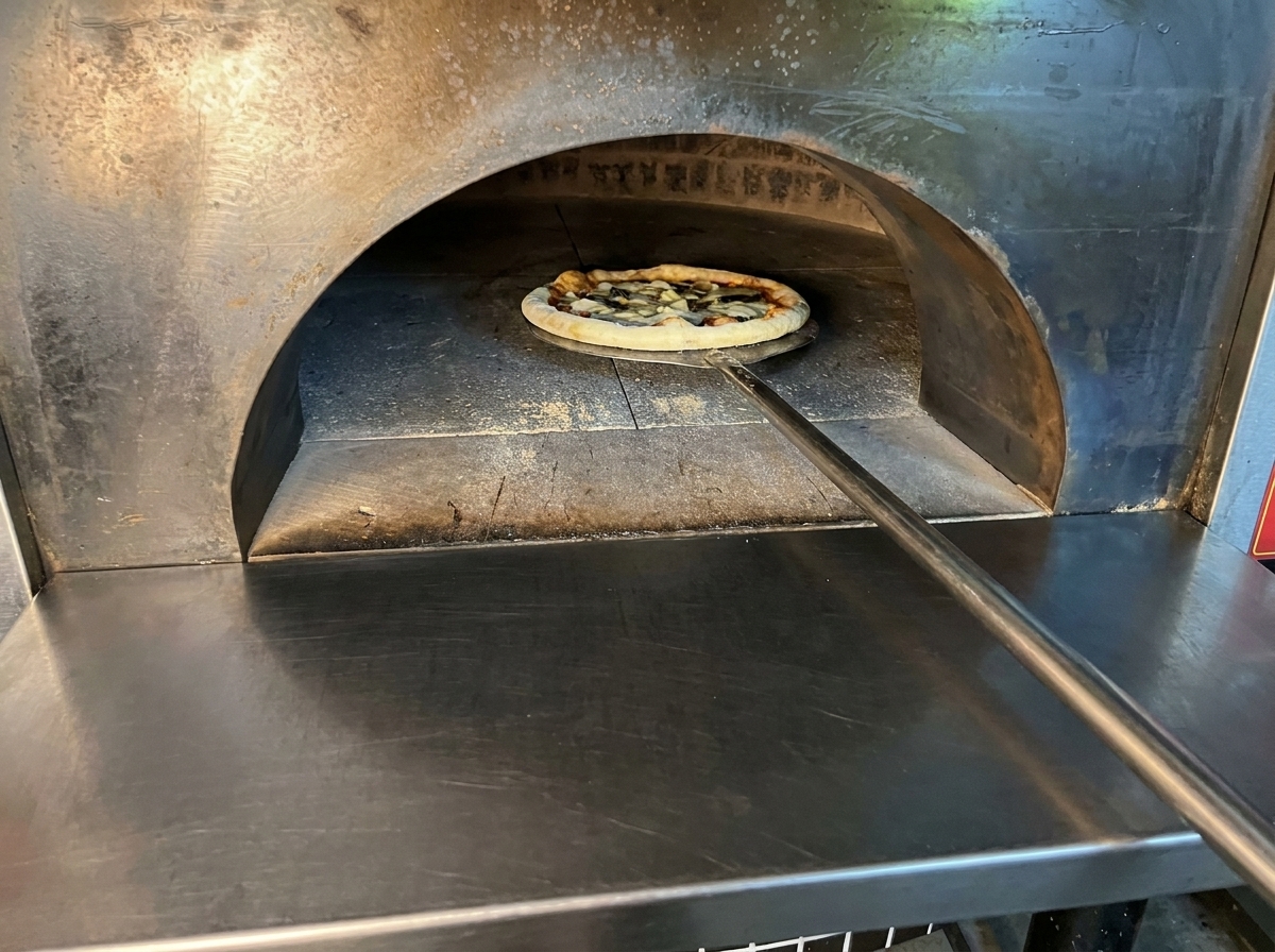 Pizza assemblée en train dêtre enfournée dans un four à bois à laide dune pelle métallique.