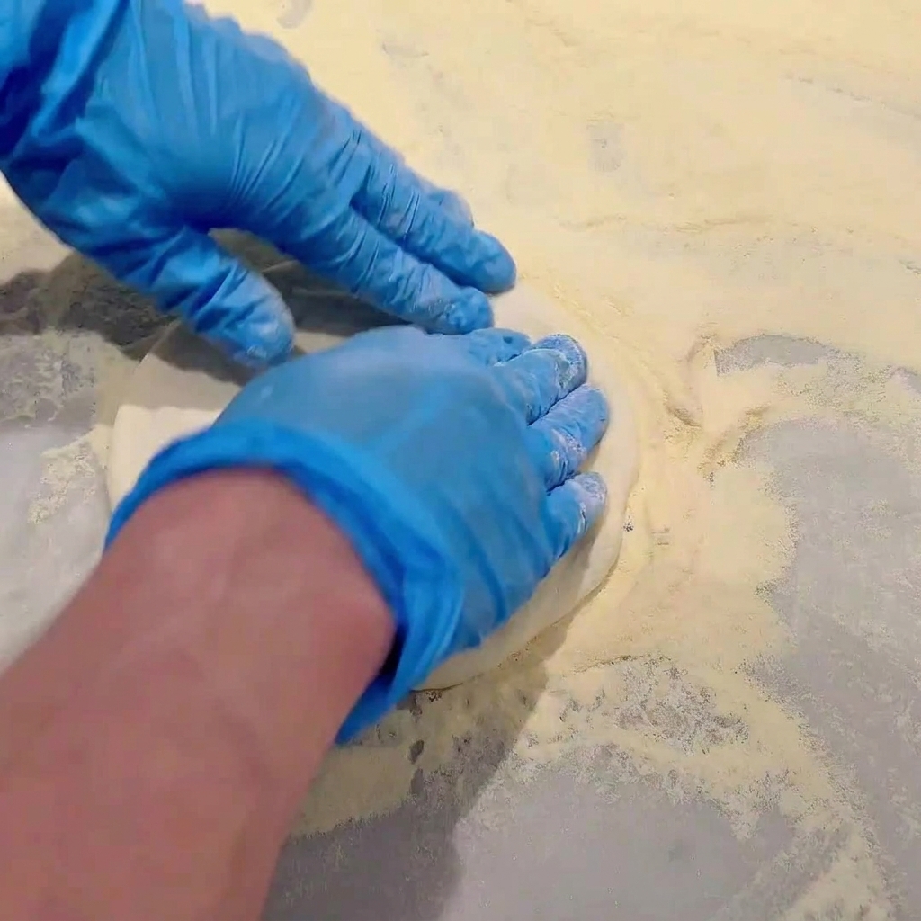 Mains gantées de bleu pressant et étirant la pâte à pizza crue sur un comptoir fariné.