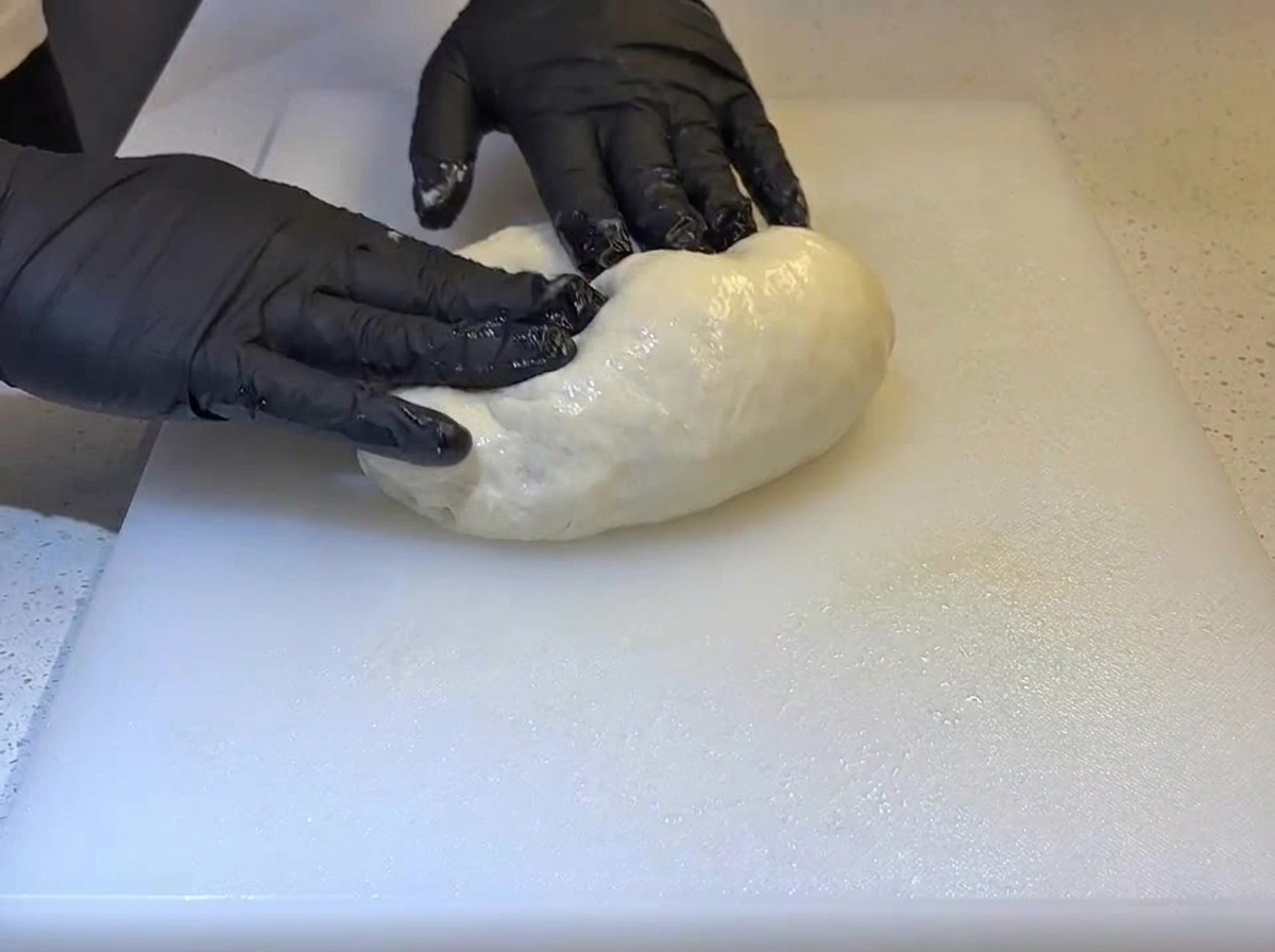 Des mains gantées de noir pliant une boule de pâte molle et huilée sur une planche à découper blanche.