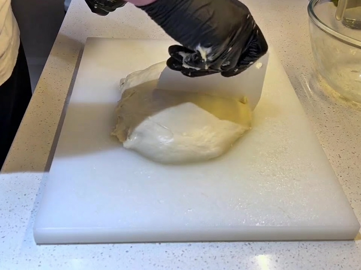 Des mains avec des gants noirs utilisant un coupe-pâte en plastique blanc pour couper un grand pâton en deux.