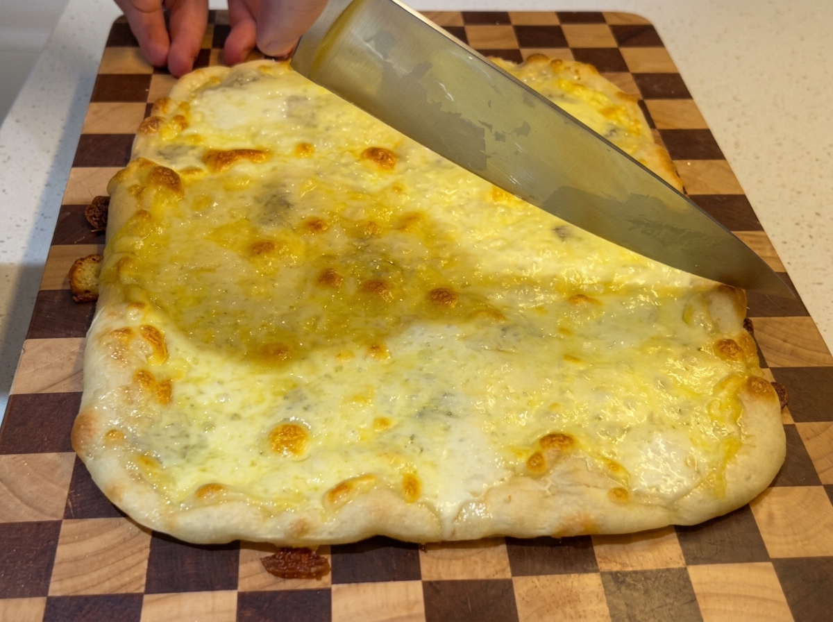 Un grand couteau de chef tranchant une pizza aux quatre fromages rectangulaire, fraîchement sortie du four, sur une planche à découper à damier.