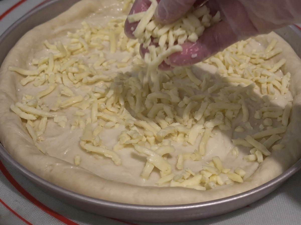 Une main gantée saupoudrant généreusement du fromage râpé sur la pâte recouverte de sauce.