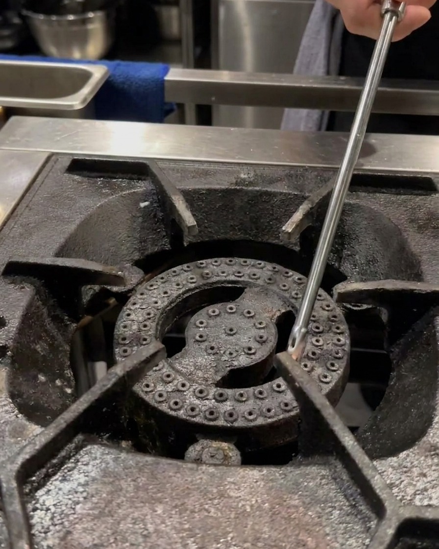 Une main utilisant un long briquet pour allumer le brûleur dune gazinière professionnelle avec une petite casserole vide posée sur la grille.