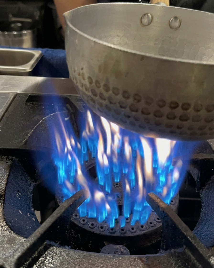 Du bouillon dos de porc versé dans une petite casserole posée sur une flamme de gaz bleue active.
