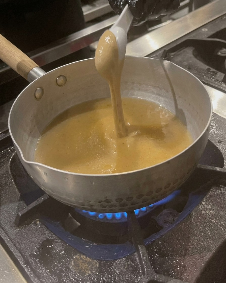 Une cuillerée de pâte de sésame épaisse de couleur beige étant mélangée dans le bouillon frémissant dans la casserole.