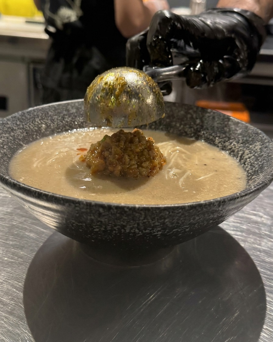 Une main gantée versant de lhuile pimentée rouge vif en cercles concentriques à la surface dun bol de ramen.