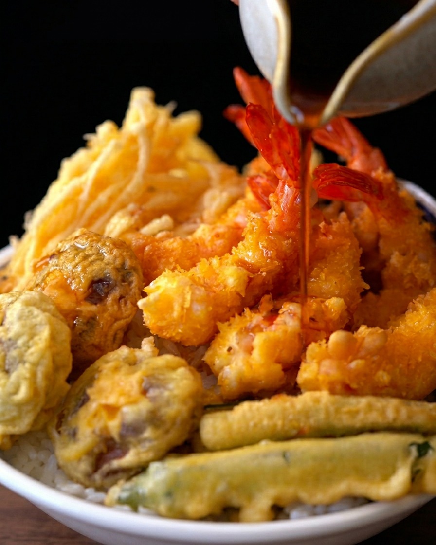 Une sauce brune et savoureuse versée depuis une verseuse en céramique sur un bol de riz surmonté de crevettes et légumes frits.