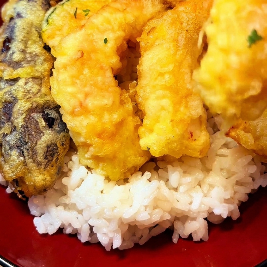 Morceaux de tempura frits dorés, incluant crevettes et légumes, déposés sur un lit de riz blanc dans un bol rouge.