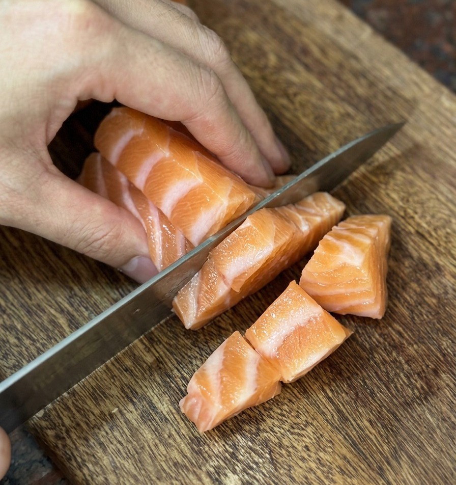 Un couteau bien aiguisé coupant du saumon frais en dés réguliers sur une planche en bois.