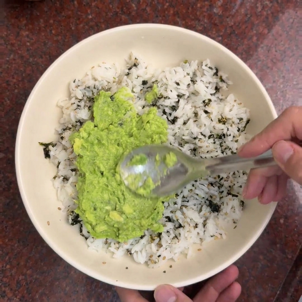 Une cuillère étalant de lavocat écrasé bien vert sur un lit de riz parsemé dalgues séchées.
