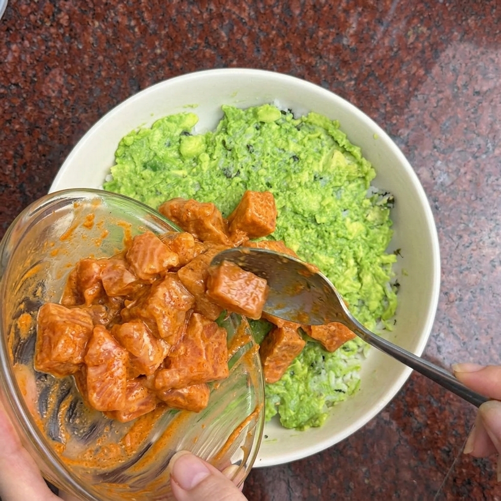 Une main déposant des morceaux de saumon en sauce sur la base de riz et davocat.