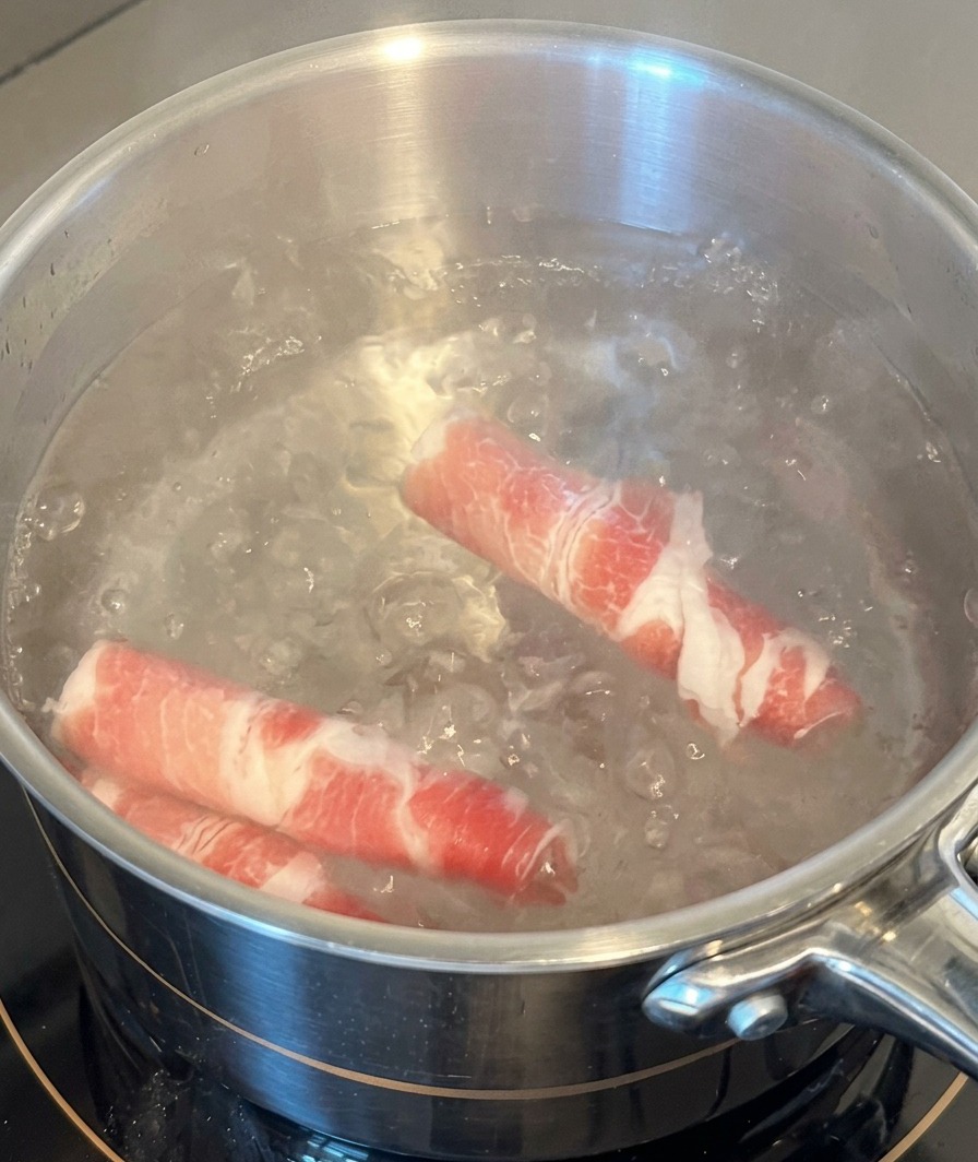 Deux rouleaux de tranches de bœuf persillé cru plongés dans une casserole en acier inoxydable deau bouillante.