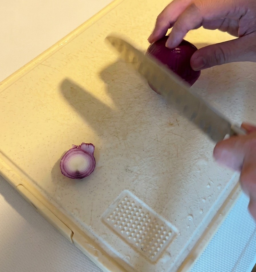 Une personne utilisant un couteau de chef pour émincer un oignon rouge entier en fines lamelles sur une planche à découper blanche.