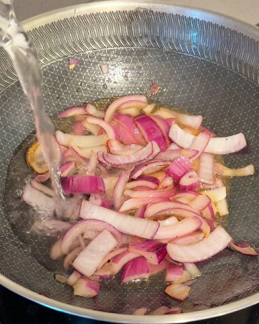 Un filet deau claire ajouté dans une poêle contenant des oignons rouges sautés.