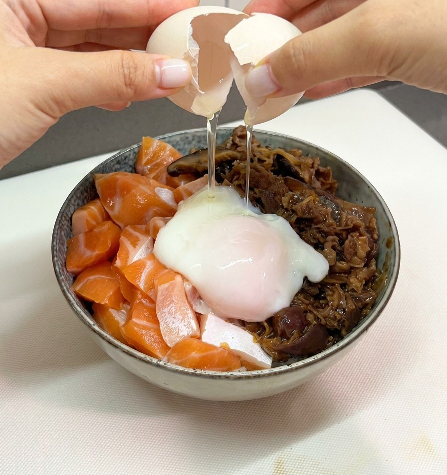 Deux mains ouvrant délicatement la coquille dun œuf blanc pour libérer un œuf onsen tremblotant sur un bol de riz au saumon et au bœuf.