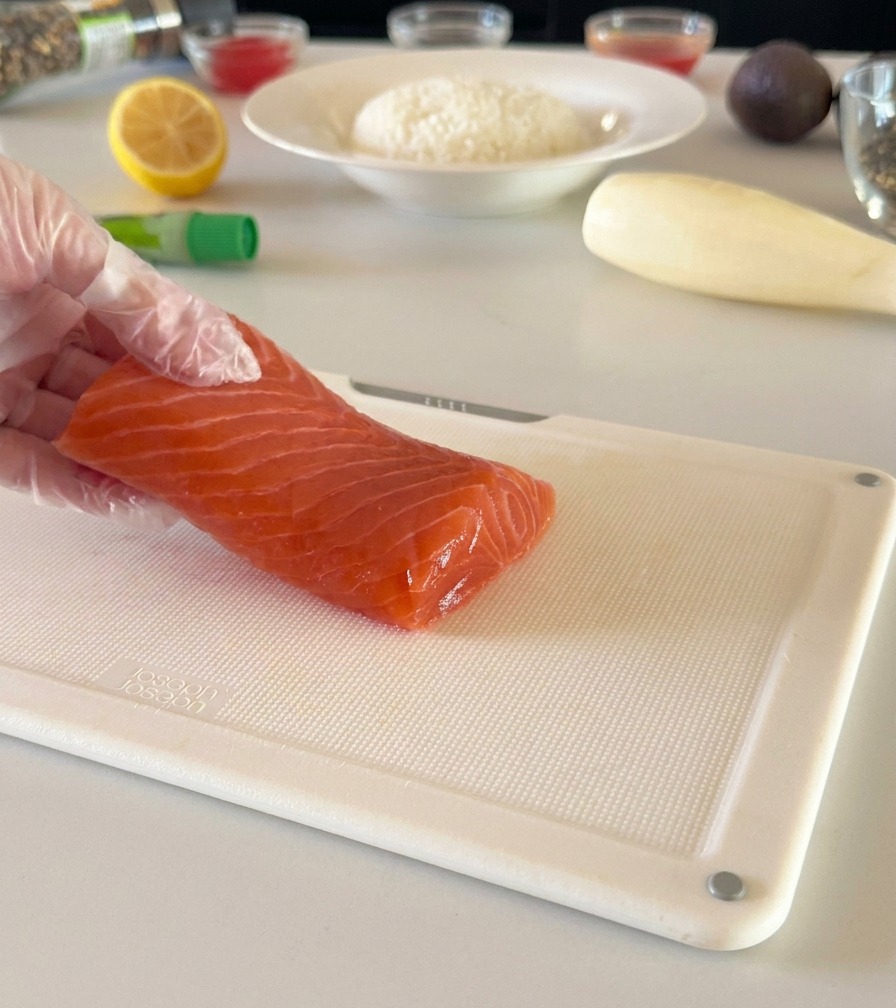 Une main gantée maintenant un filet de saumon frais sur une planche à découper blanche, prête à le couper en dés.