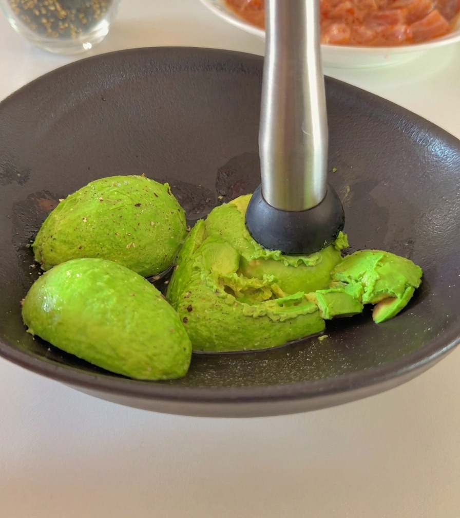 Une personne utilisant un presse-purée en métal pour écraser des moitiés davocat mûr dans un bol en céramique sombre.