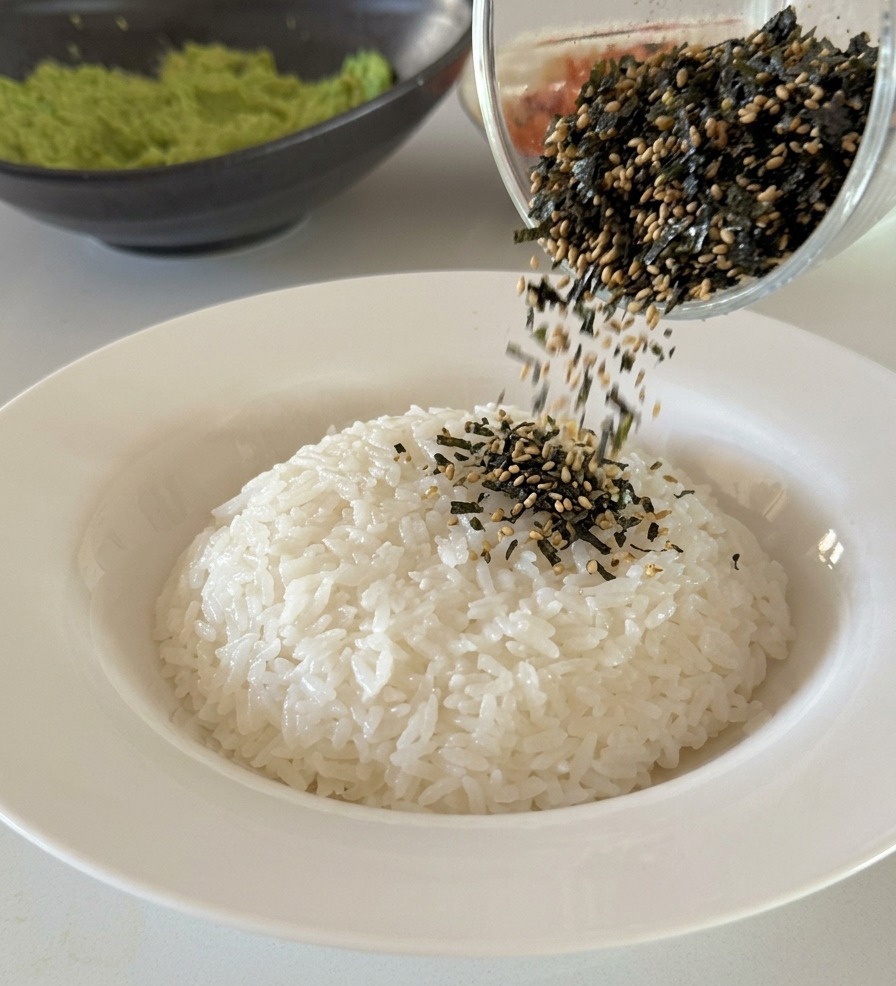 Un mélange de lanières dalgues noires et de graines de sésame blanches versé depuis un verre sur un dôme de riz blanc.