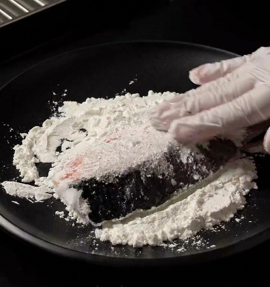 Une main gantée pressant un pavé de saumon frais dans de la fécule blanche sur une assiette noire pour lenrober uniformément.