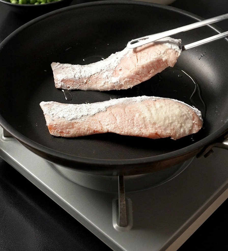 Une pince de cuisine déposant un pavé de saumon enrobé de fécule dans une poêle chaude à côté dun autre pavé.