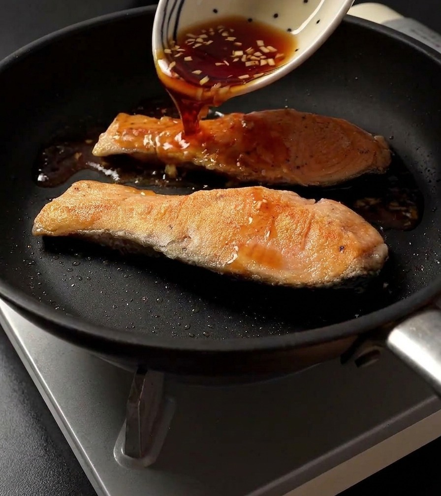Versement dune sauce teriyaki foncée depuis un bol blanc à motifs sur deux pavés de saumon saisis dans une poêle.