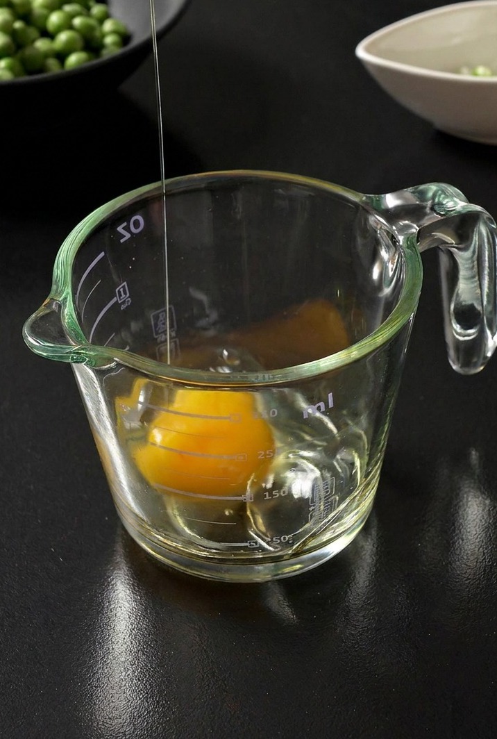 Un œuf frais cassé dans un verre doseur en verre transparent, avec un bol de petits pois en arrière-plan flou.