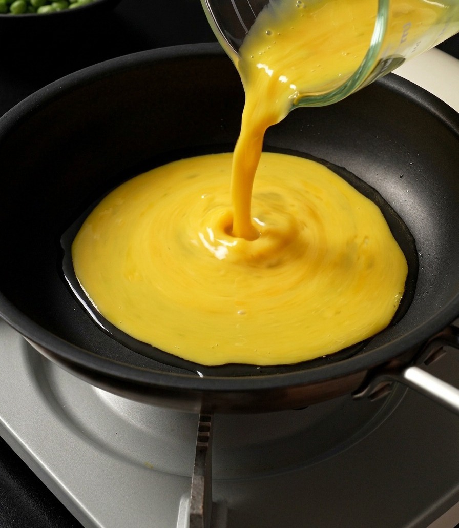 Le mélange dœufs battus jaunes versé depuis un verre doseur dans une grande poêle pour former une omelette très fine.