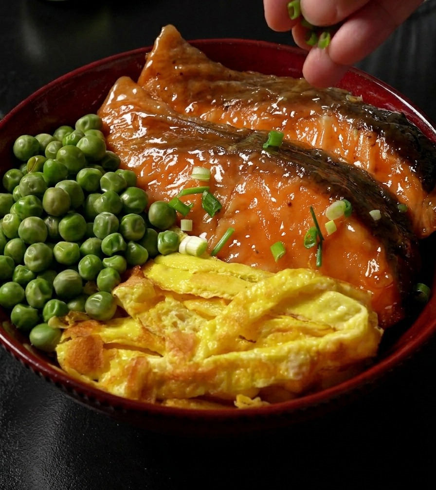 Une main parsemant des oignons verts émincés sur un bol de riz terminé, surmonté de saumon teriyaki laqué, de lanières dœuf et de petits pois.