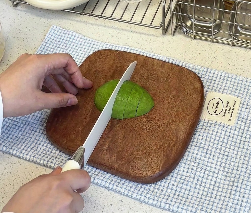 Une hand maintenant une moitié davocat pelé sur une planche en bois pendant quun couteau blanc la coupe en petits cubes.