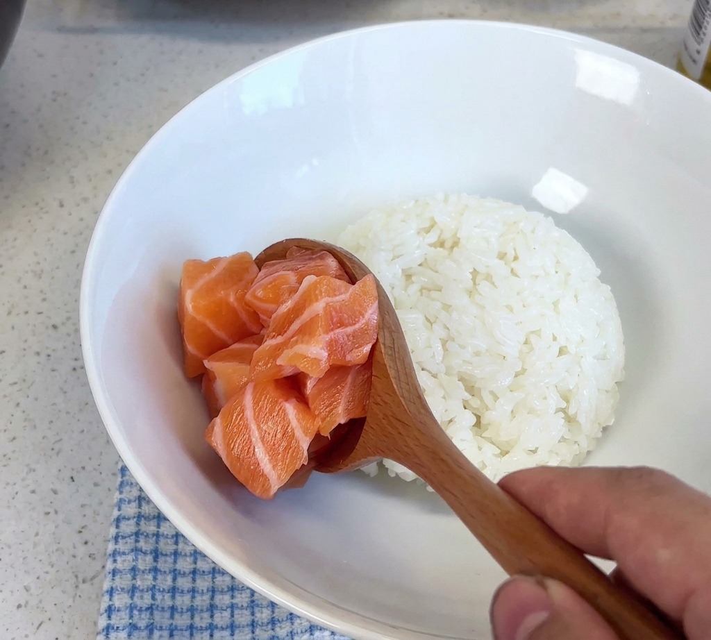 Une cuillère en bois déposant une portion de saumon cru en dés dans un bol blanc contenant du riz cuit.