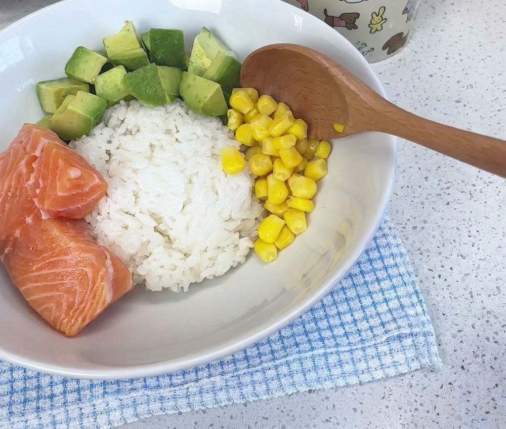 Une cuillère en bois ajoutant des grains de maïs jaune vif dans un bol blanc garni de riz, davocat et de saumon.