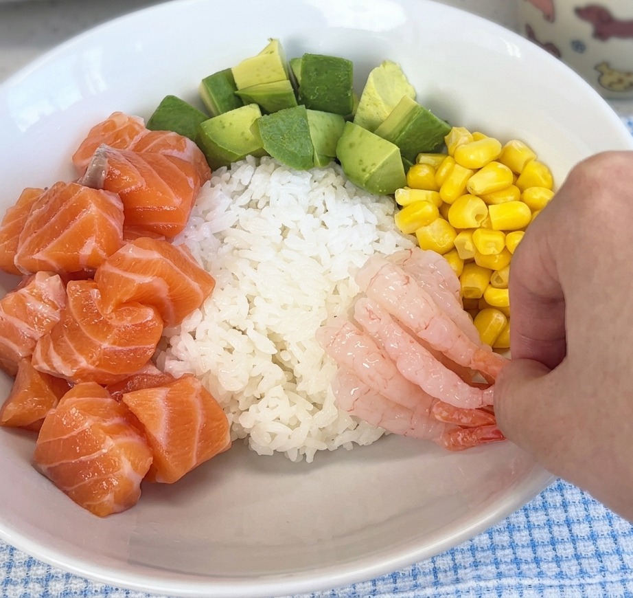 Une main déposant des crevettes douces roses et translucides sur le riz, aux côtés du saumon et du maïs.