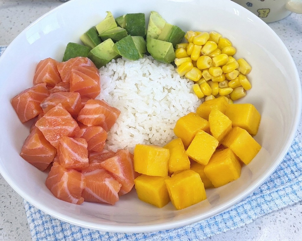 Un poke bowl coloré garni de riz, saumon, avocat, maïs et crevettes, complété par une portion de dés de mangue jaune vif.