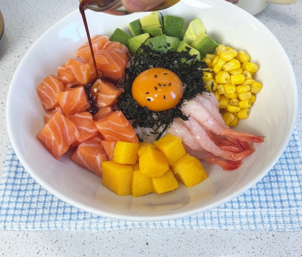 Sauce soja sombre au wasabi versée depuis un petit récipient en verre sur le poke bowl de saumon assemblé.