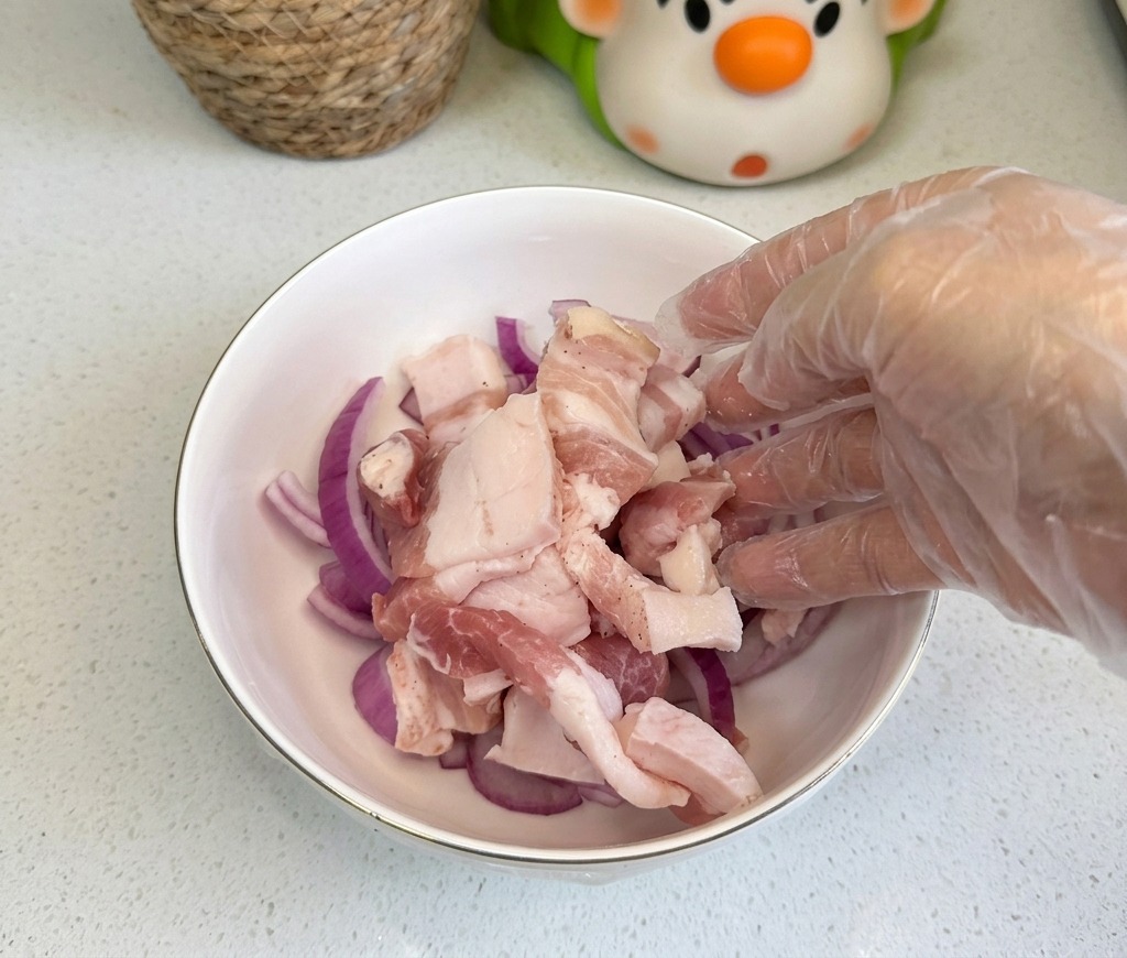 Tranches de poitrine de porc crue ajoutées dans un bol contenant des oignons rouges émincés.