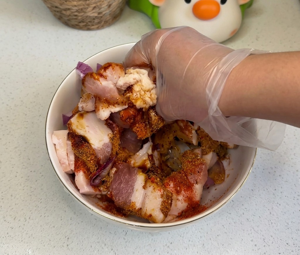 Une main gantée mélangeant soigneusement la poitrine de porc avec les oignons et les épices colorées.