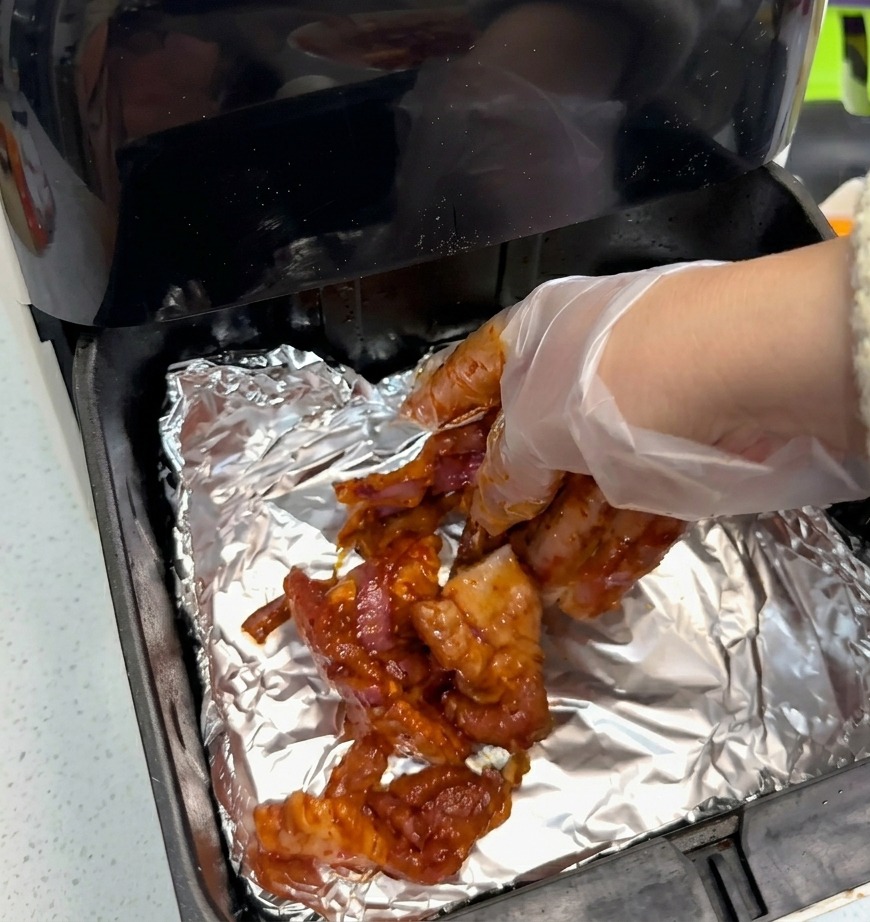 Une main gantée déposant des tranches de porc mariné sur du papier daluminium dans le panier du air fryer.