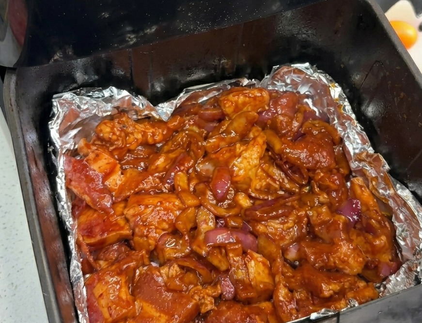 La poitrine de porc marinée et les oignons répartis uniformément au fond du panier du air fryer doublé daluminium.