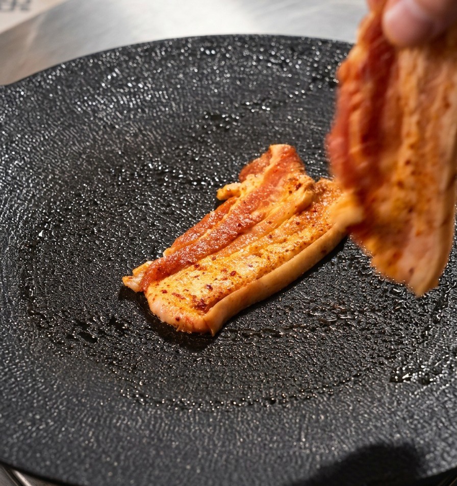 Une main déposant une tranche de poitrine de porc sur une plaque de gril ronde et noire.