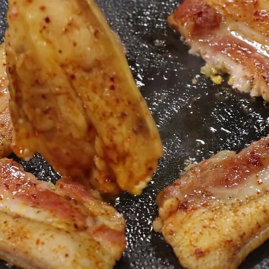 Des pinces métalliques retournant un morceau de poitrine de porc doré sur un gril brûlant.
