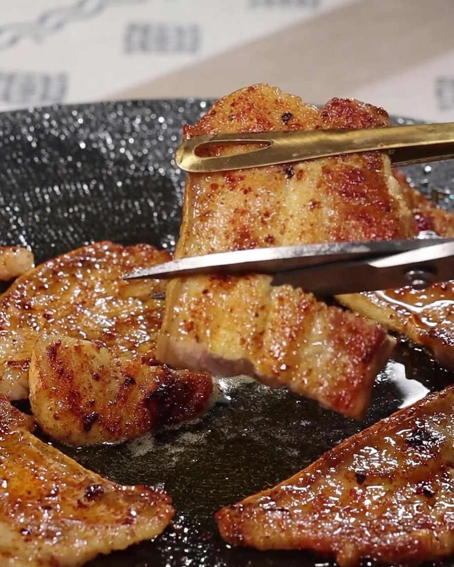 Des ciseaux de cuisine en métal coupant un morceau de poitrine de porc grillé au-dessus de la poêle.