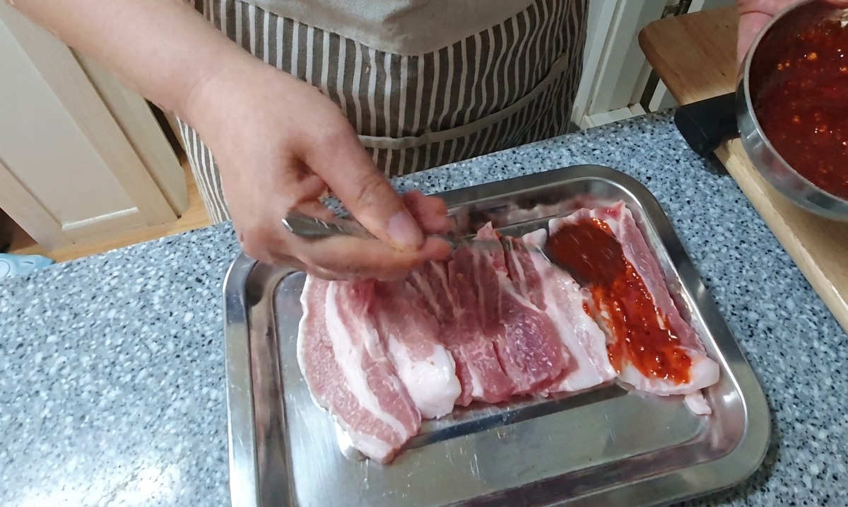 Une main badigeonne des tranches de poitrine de porc crue avec une épaisse marinade rouge au piment sur un plateau argenté.