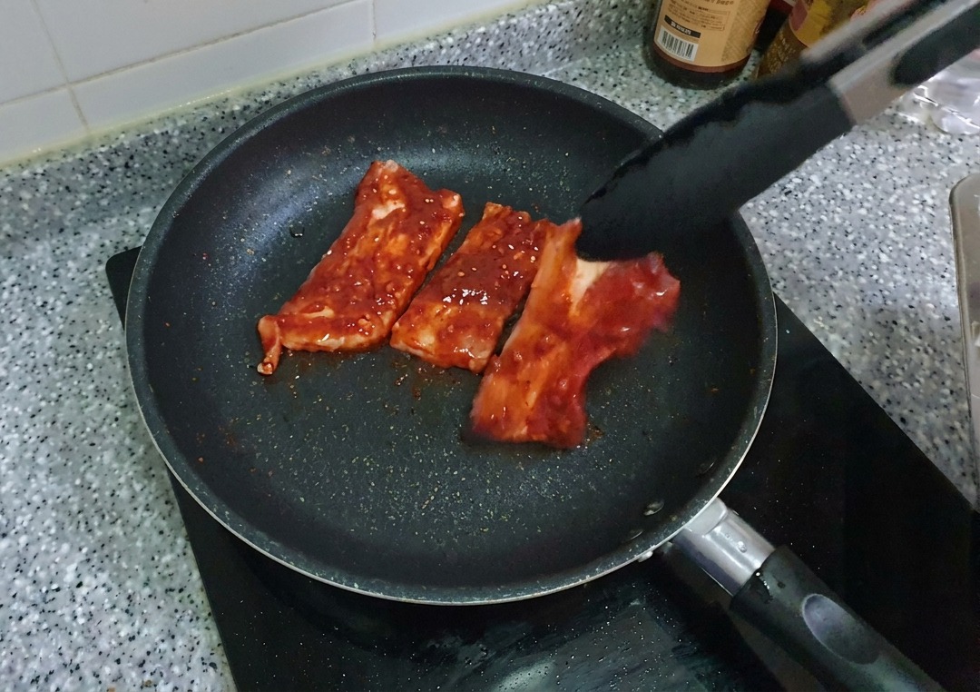 Tranches de poitrine de porc rouge marination déposées dans une poêle à frire antiadhésive noire à laide dune pince.
