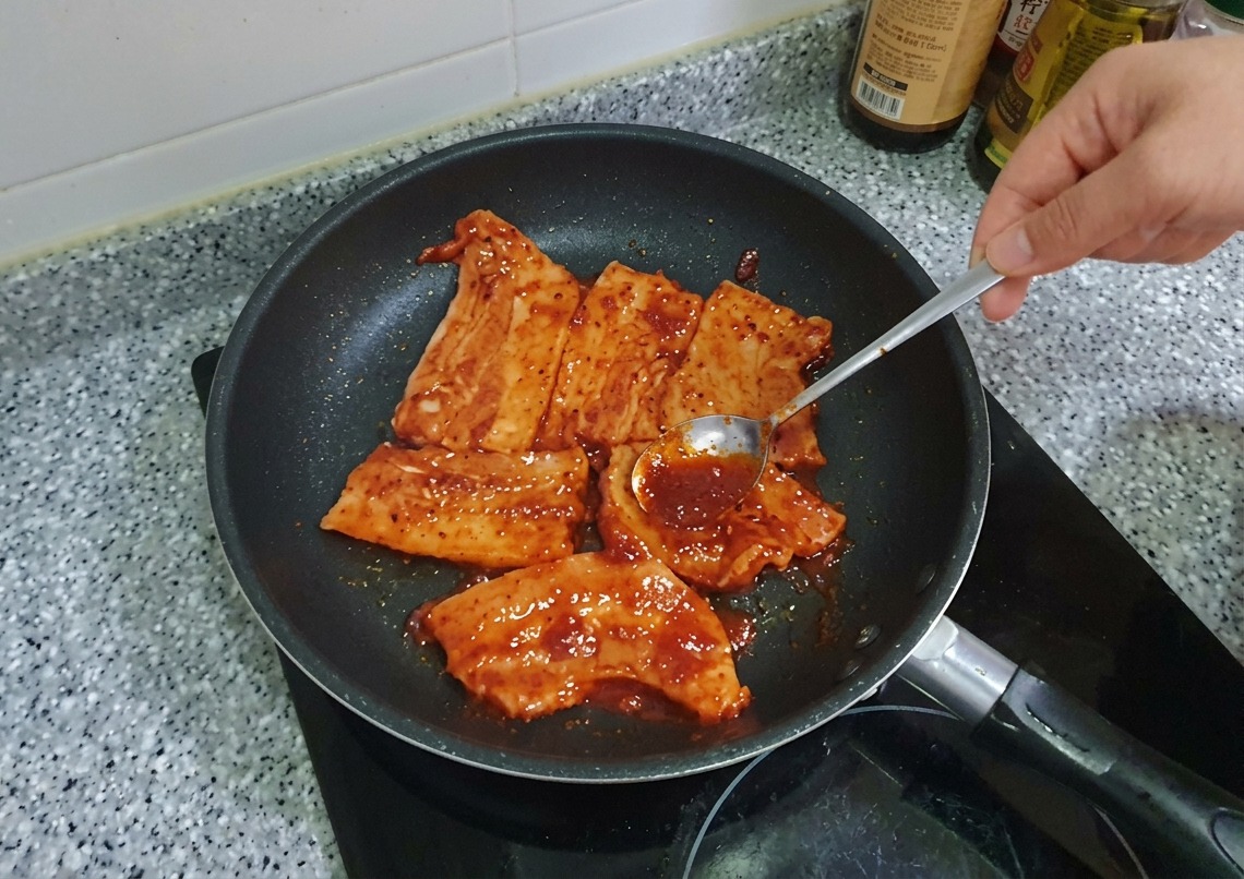 Une cuillère étale une épaisse marinade rouge au Gochujang sur des tranches de poitrine de porc dans une poêle.