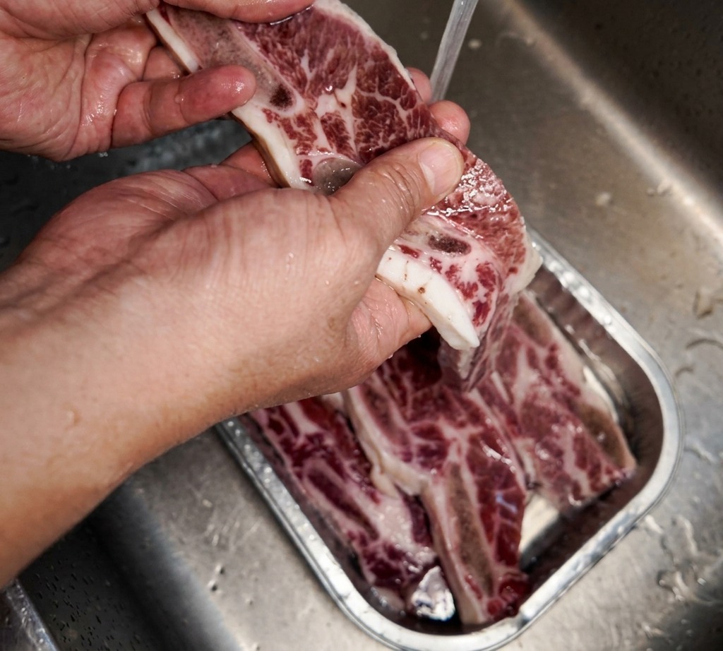 Une main tenant un morceau de travers de bœuf persillé sous un filet deau froide au-dessus dun évier en inox.