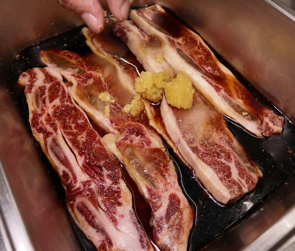 Une main déposant un petit tas de gingembre fraîchement haché sur des travers de bœuf en train de mariner dans un plateau en inox.