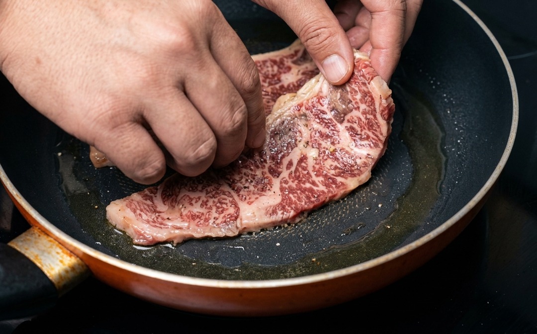 Une main déposant un travers de bœuf mariné et bien persillé dans une poêle chaude.